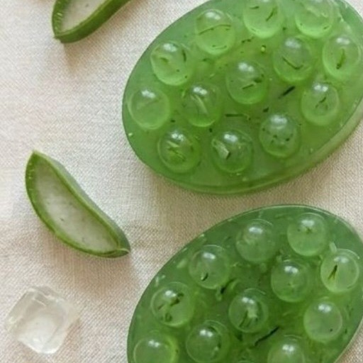 Dos jabones artesanales de aloe vera, verdes y translúcidos, rodeados de hojas de aloe y cubos de hielo sobre lino