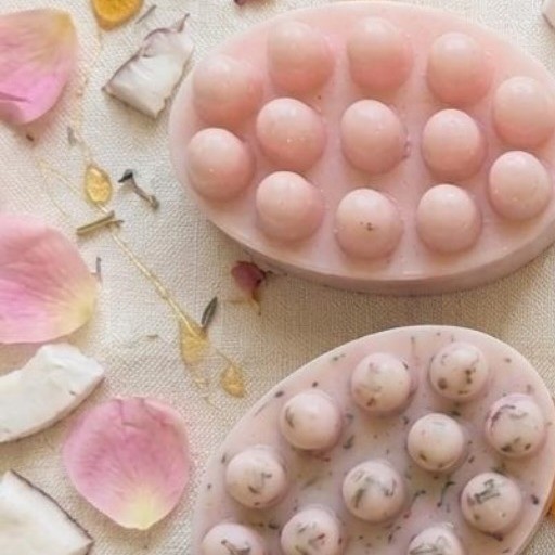 Dos jabones artesanales rosados, entre pétalos de rosa, trozos de coco y gotas de miel sobre lino